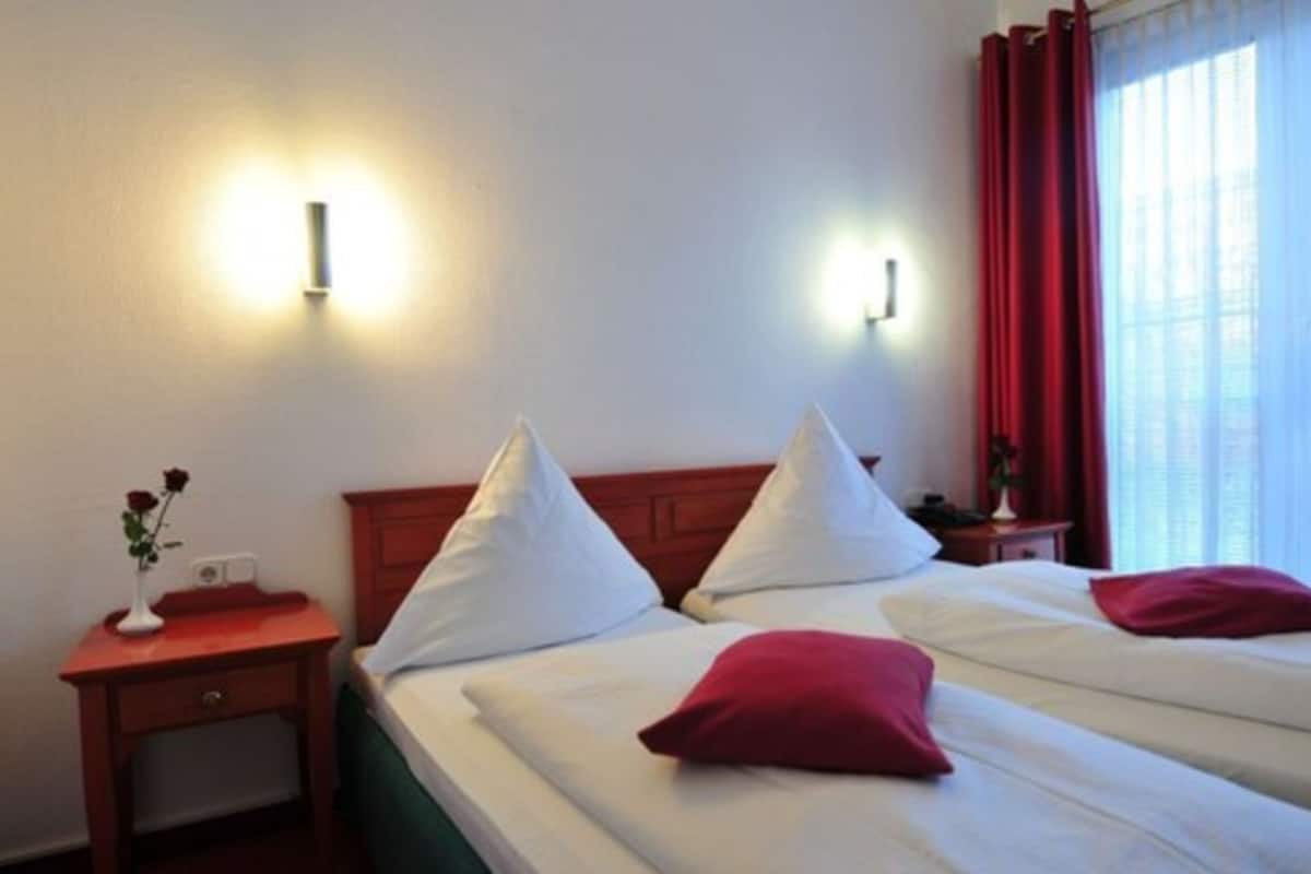 Une chambre d’hôtel avec deux lits, chacun agrémenté de draps blancs et d’oreillers rouges, une table de chevet en bois ornée d’un vase de fleurs et des lampes fixées au mur.