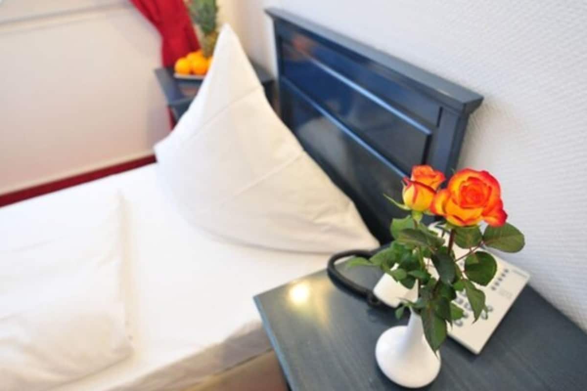 Un lit avec une literie blanche, un oreiller blanc et une table de chevet sur laquelle se trouve un vase blanc contenant des roses oranges.