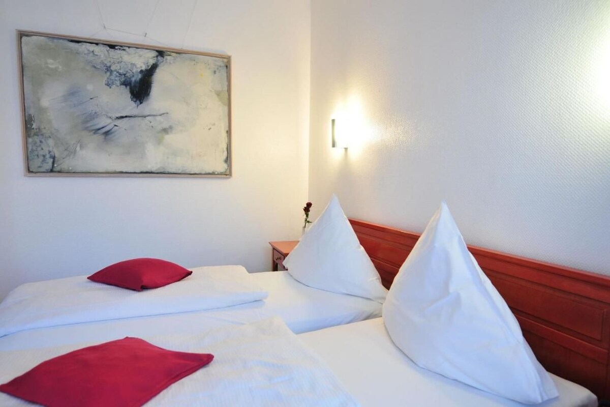 A hotel room with two beds, white bedding, red pillows, a wooden headboard, a wall-mounted abstract painting, and a bedside lamp.