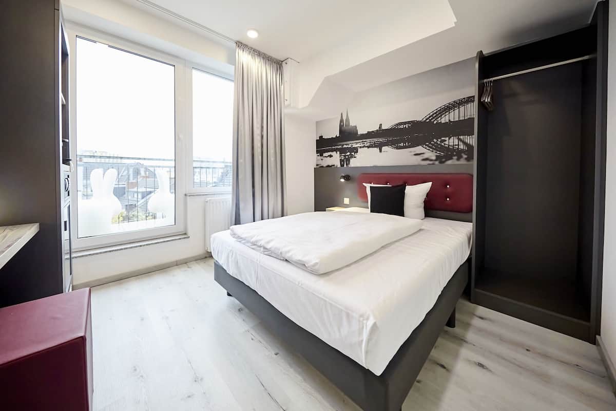 A modern hotel room with a large bed, a red headboard, a cityscape mural, and a view of a bridge.