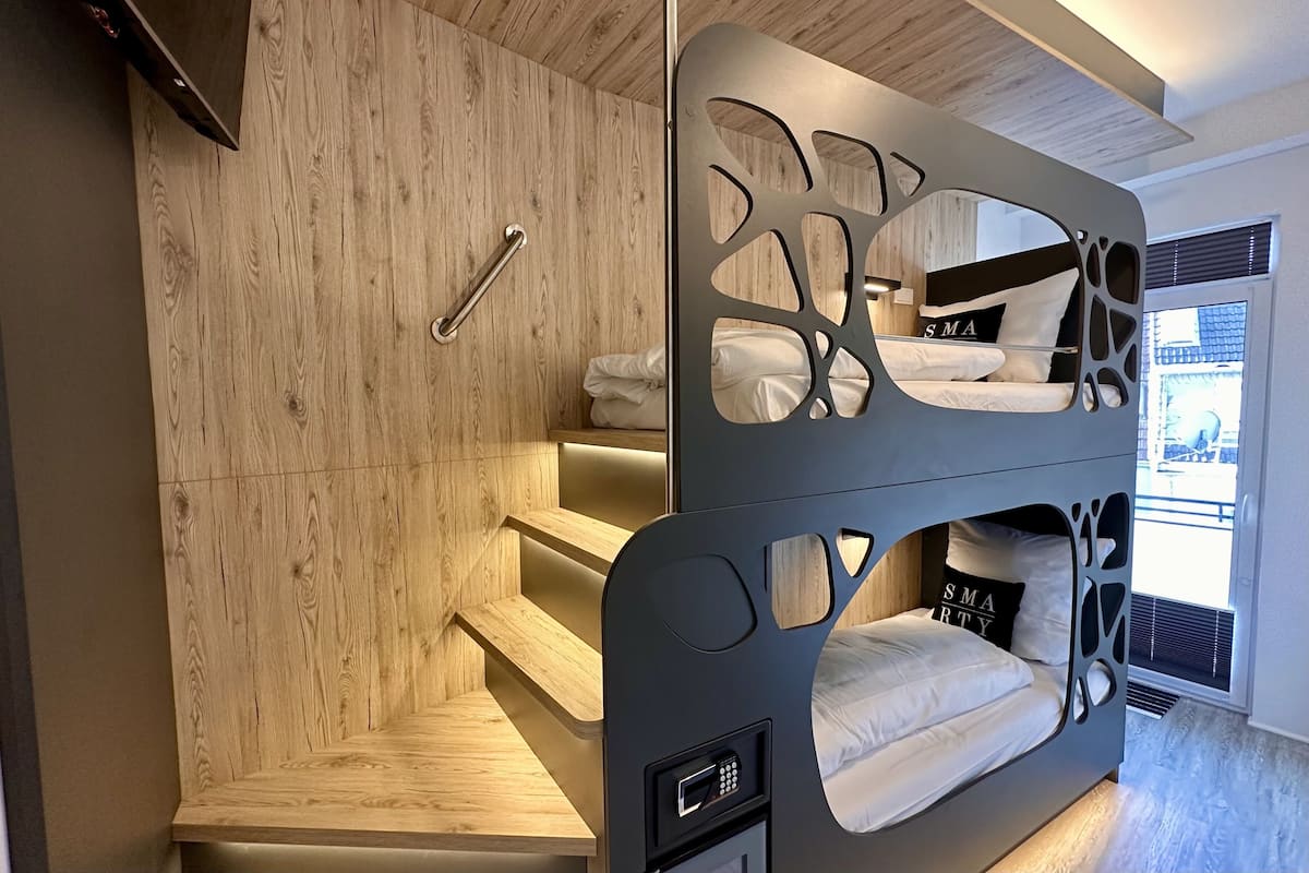 A modern hotel room with a bunk bed, wooden panel walls, a small seating area, and a window with a view.