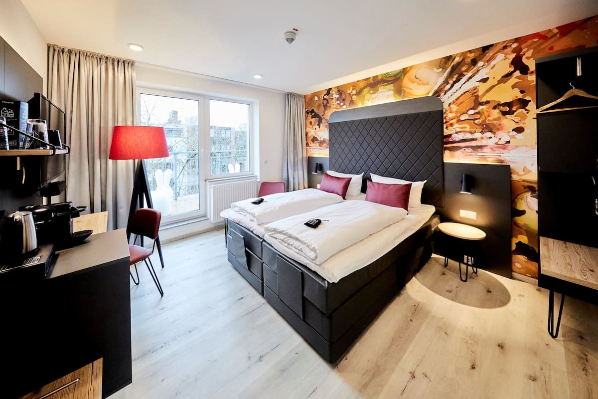 A modern hotel room with a large bed, a desk with a lamp, a chair, and a view of the city through the window.