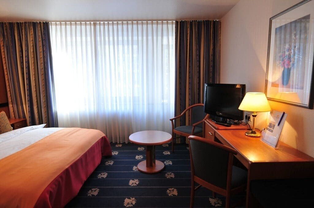 Habitació d'hotel amb un llit, una taula de nit amb televisió, una cadira, una taula petita i una làmpada.