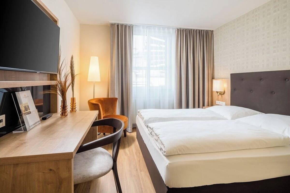 A hotel room with a bed, a desk with a chair, a TV, and a window with curtains.