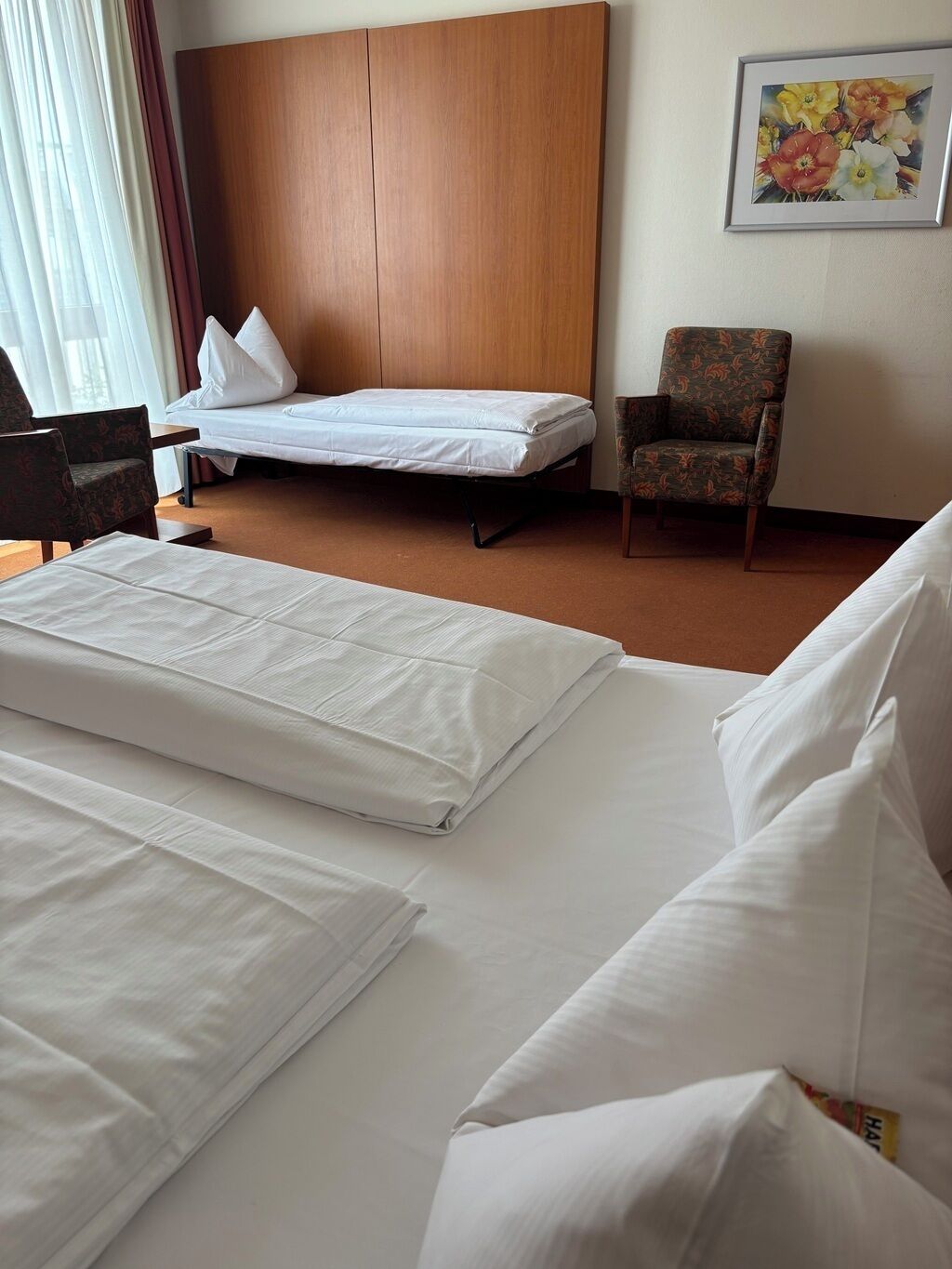 Ein Hotelzimmer mit zwei Betten, einem Holzkopfende, einem Sessel und einem Gemälde an der Wand.