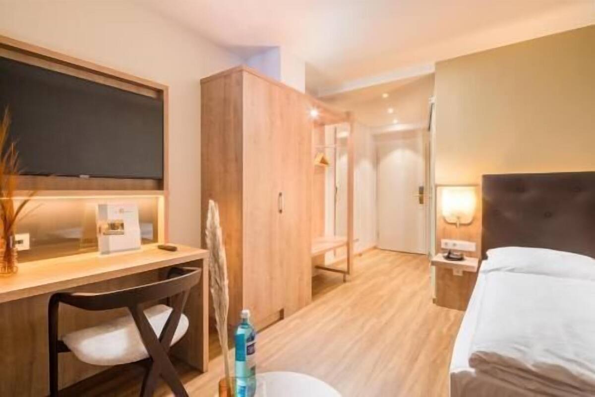 A hotel room with a bed, a desk with a chair, a wardrobe, a TV, and a bathroom visible through an open door.