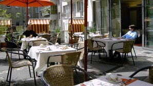 Un café al aire libre con mesas y sillas, cubierto por sombrillas naranjas, y un edificio al fondo.