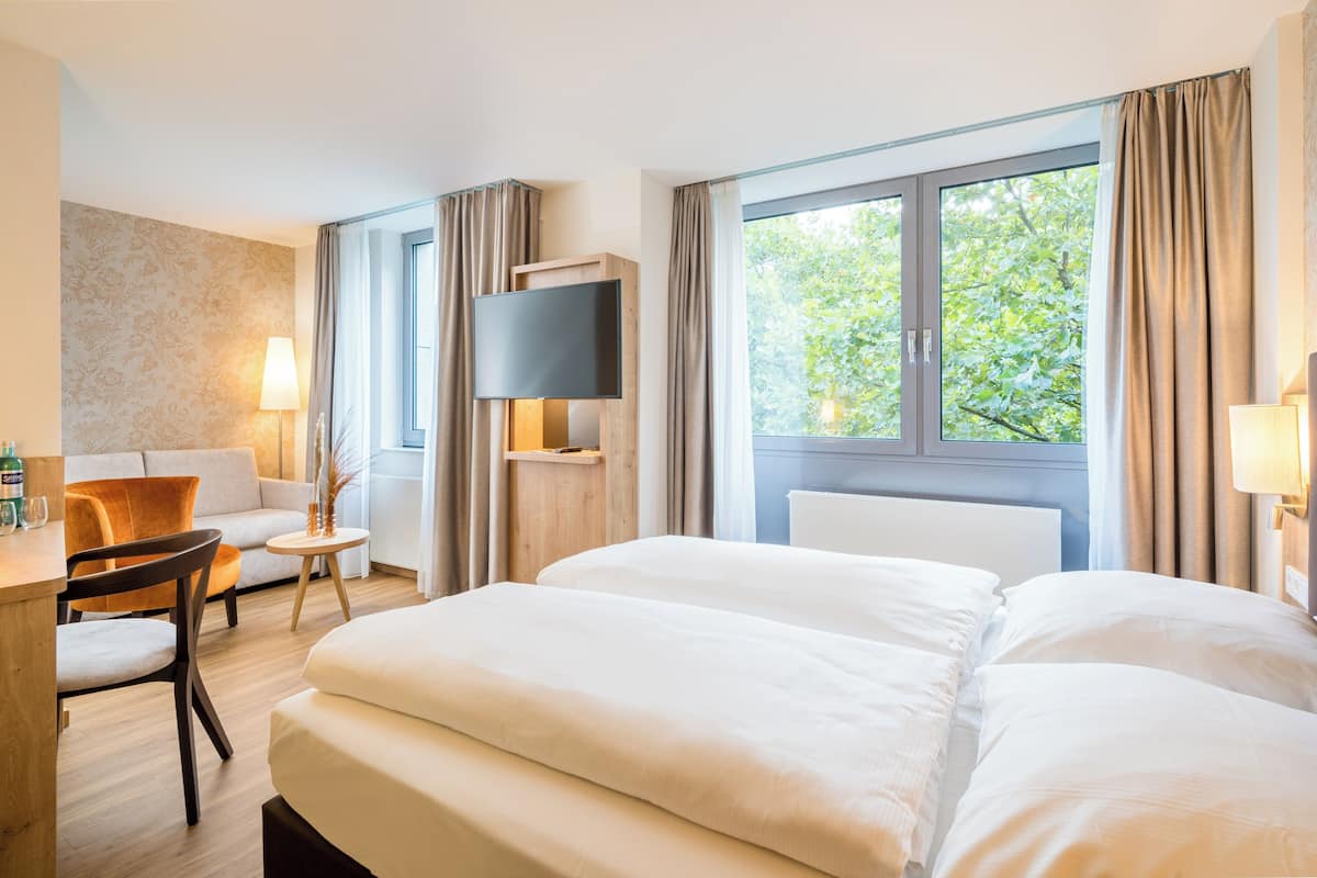 A hotel room with a large bed, a TV, a desk with a chair, and a view of greenery through the window.