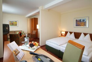 A hotel room with a bed, a desk with a remote control, a chair, a dining table with a fruit bowl, and a television.