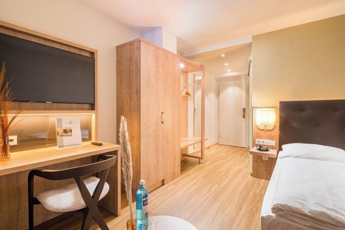 A hotel room with a wooden desk, a bed, a TV mounted on the wall, and a nightstand with a lamp.