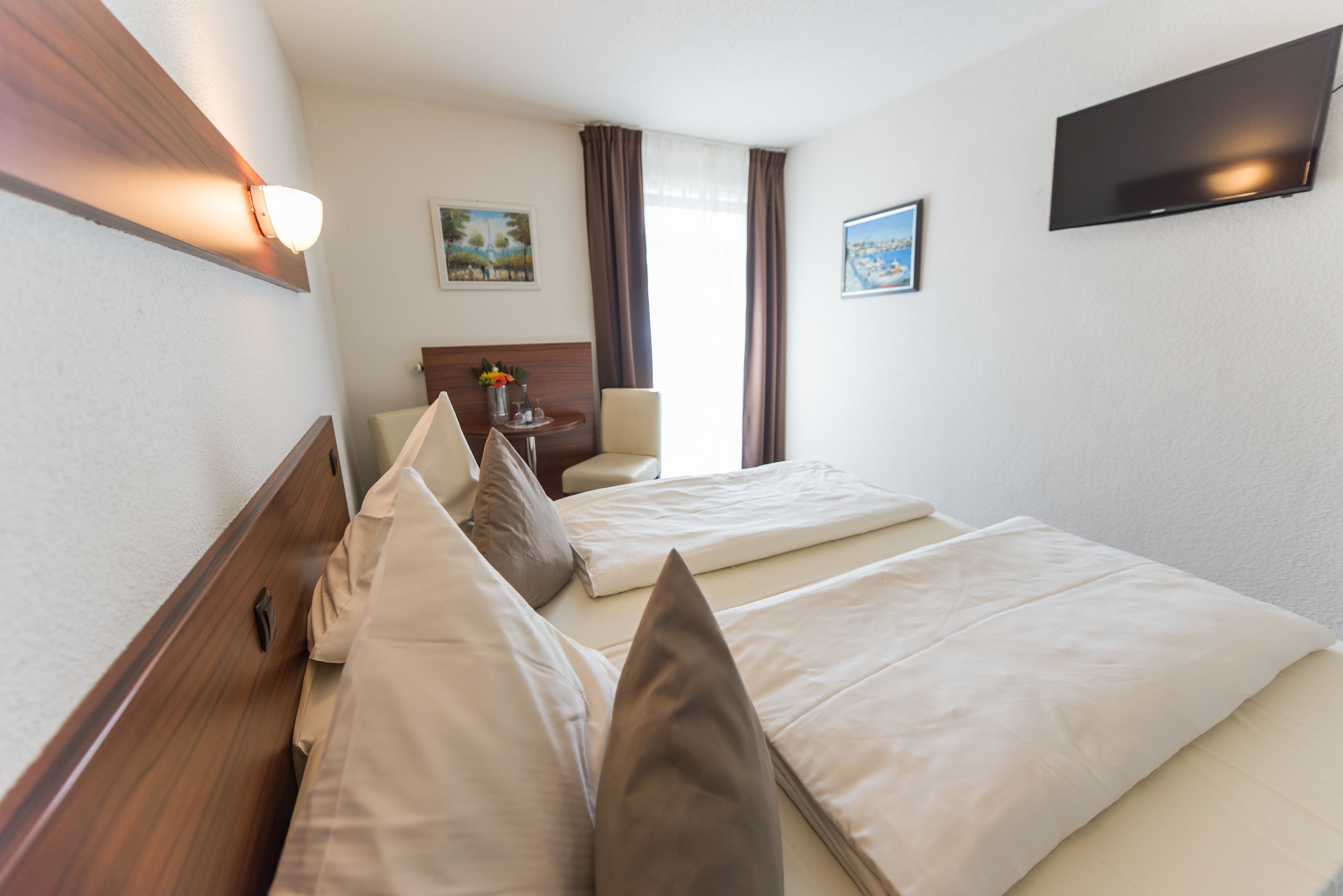 A hotel room with two beds, a TV mounted on the wall, and a small table with a vase of flowers.