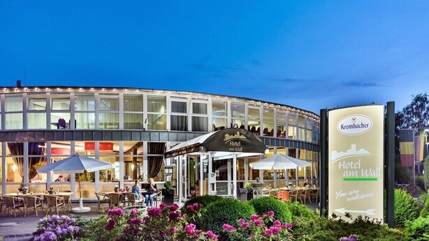 A hotel with a curved facade, outdoor seating area, and a sign welcoming guests. - Hotel Am Wall Soest (Soest)