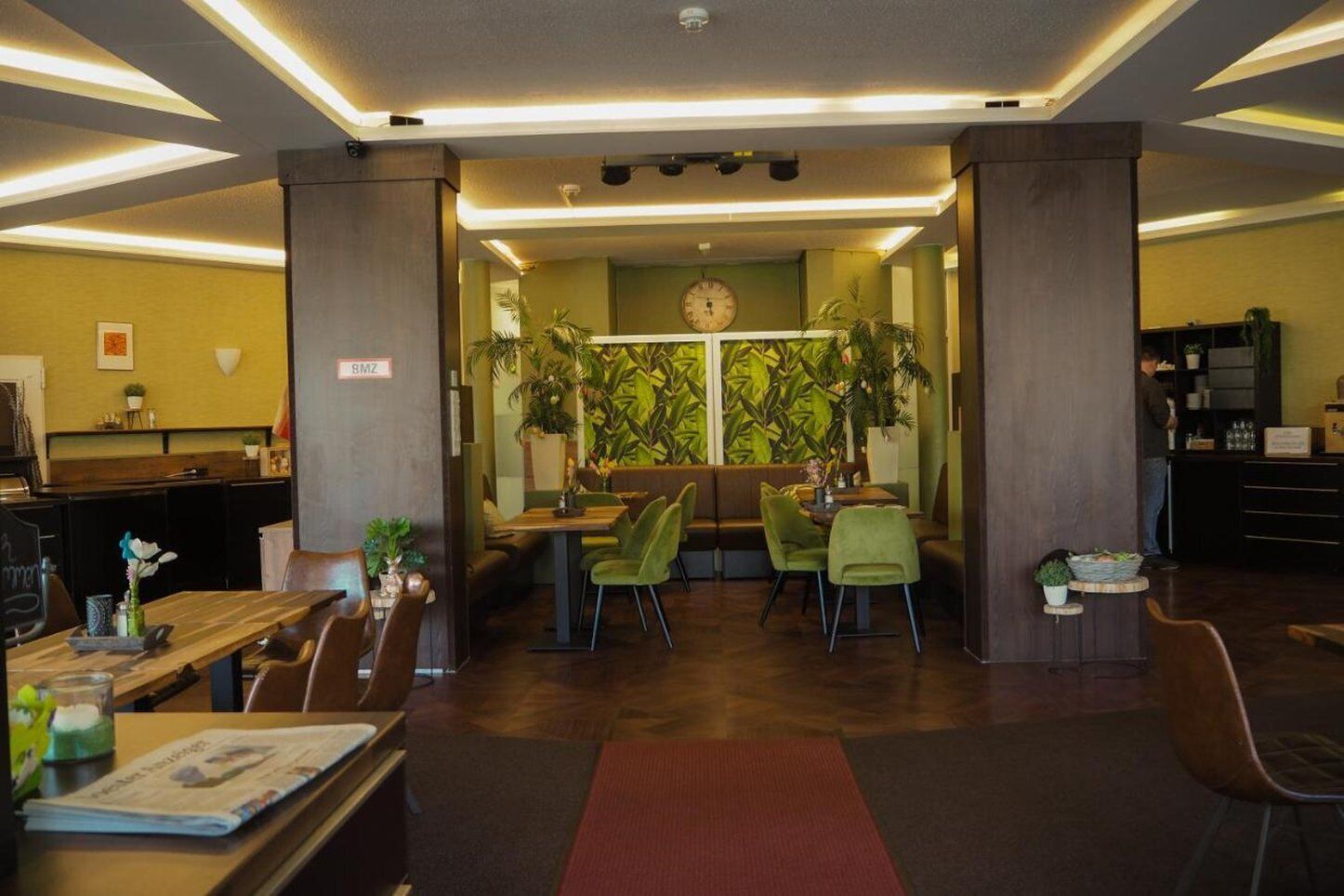 Un restaurante moderno con una zona de estar central, sillas verdes y un amplio ventanal con un reloj.