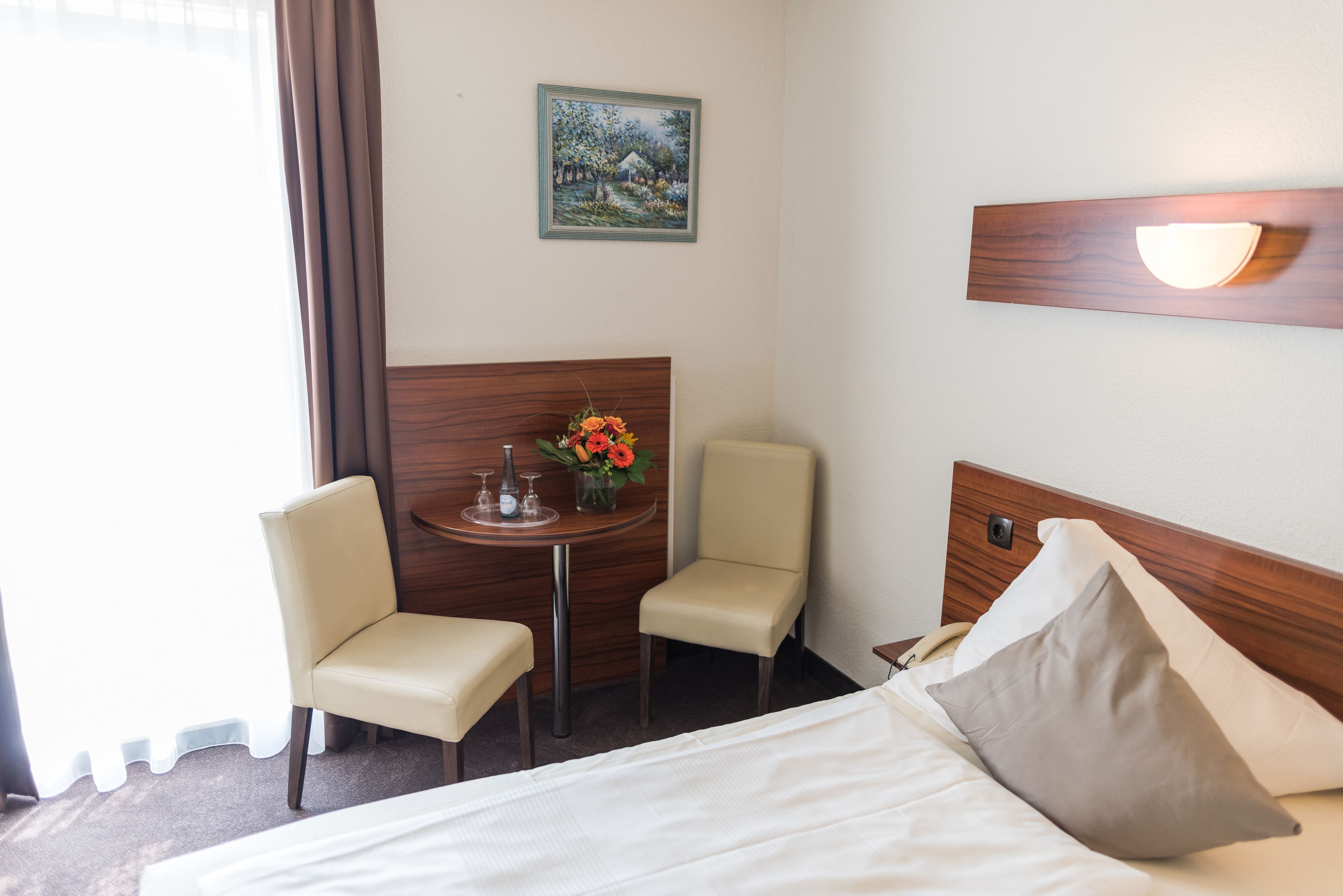 A hotel room with a bed, two chairs, a small table with a vase of flowers, a painting on the wall, and a nightstand with a lamp.