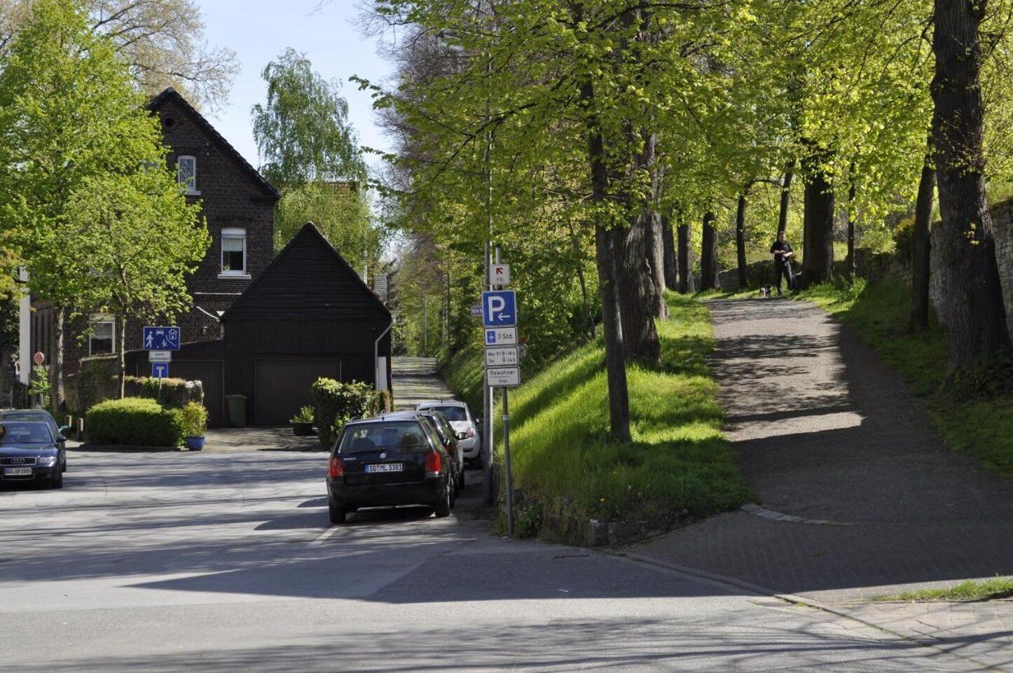 A residential street with parked cars, a pedestrian path, and a building with a dark roof.