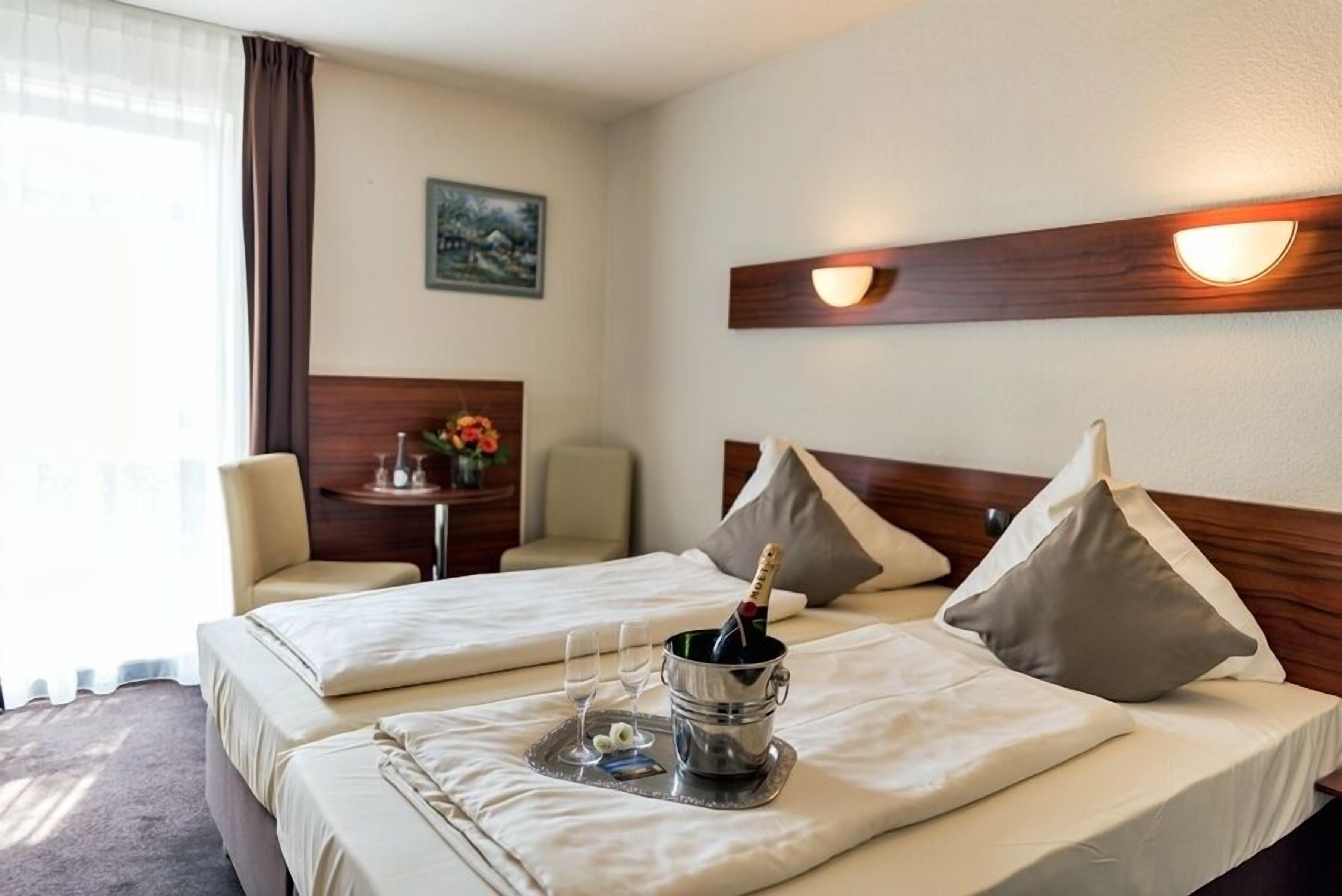 A hotel room with two beds, a bedside table with a lamp, a painting on the wall, and a window with curtains.
