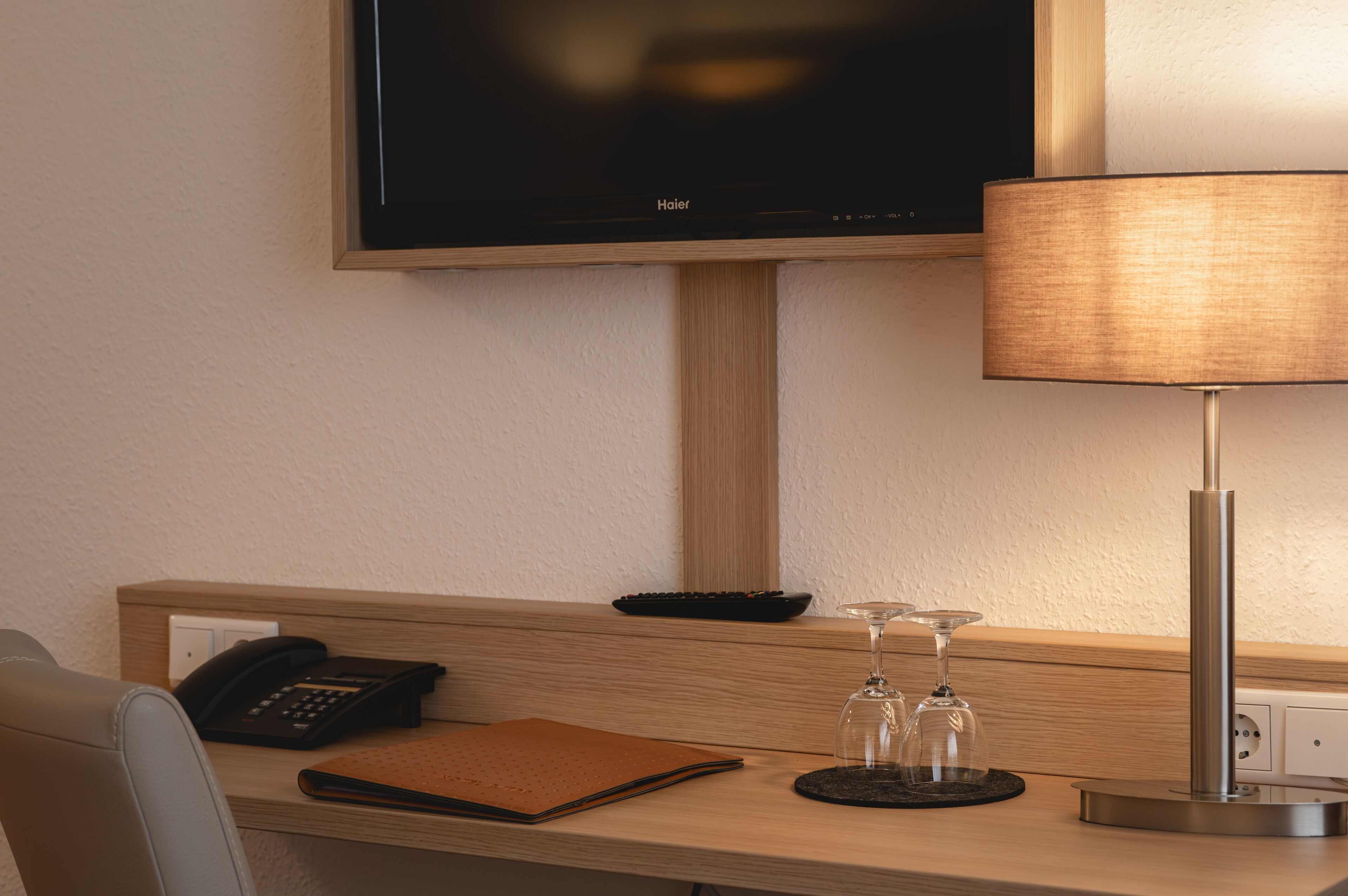 A hotel room with a wooden desk, a black telephone, a brown notebook, two wine glasses, and a lamp.