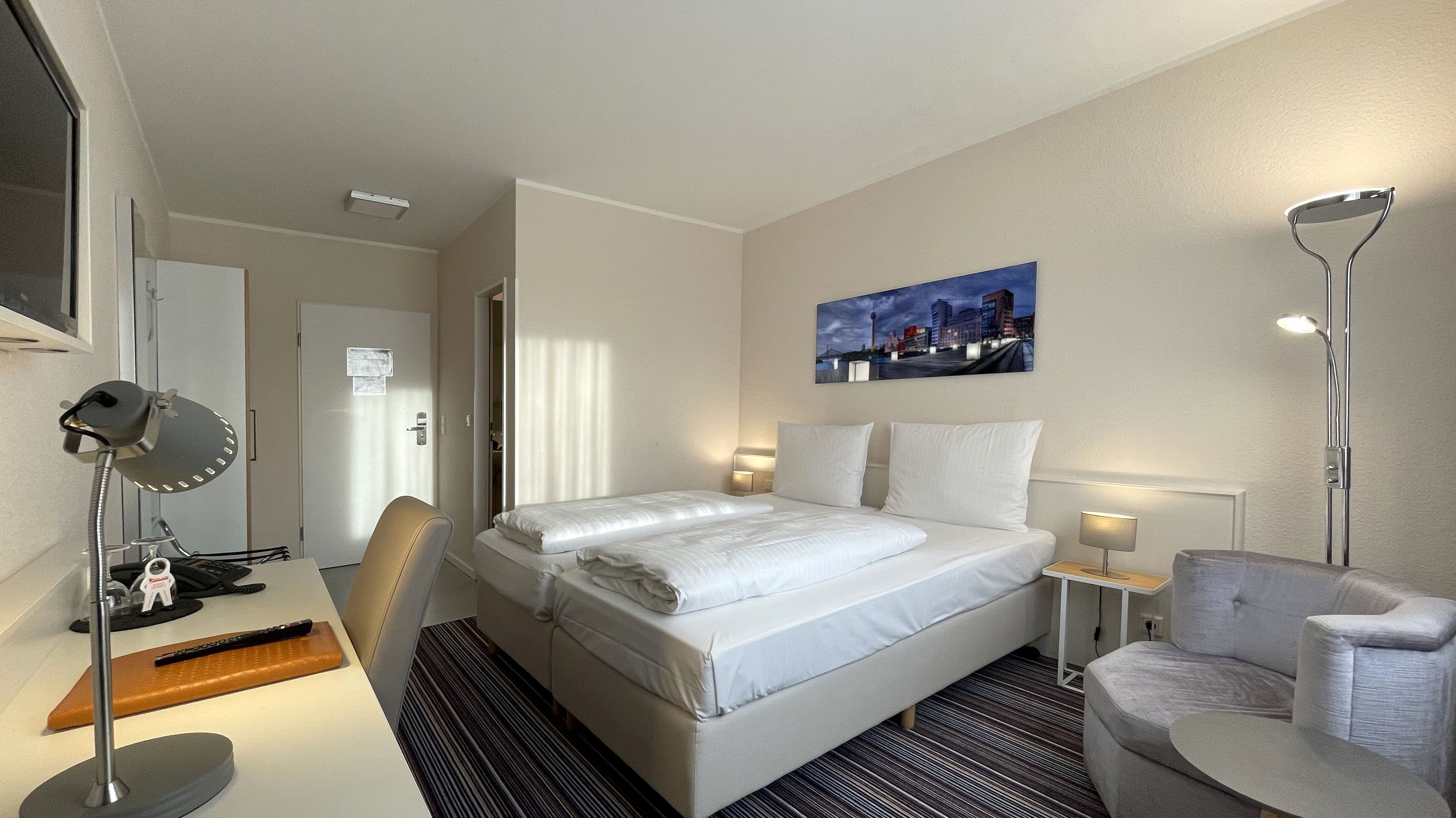 A hotel room with a bed, a desk, a chair, a standing lamp, and a painting on the wall.