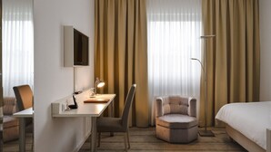 Habitación de hotel con escritorio, silla, sillón, cama y cortinas.