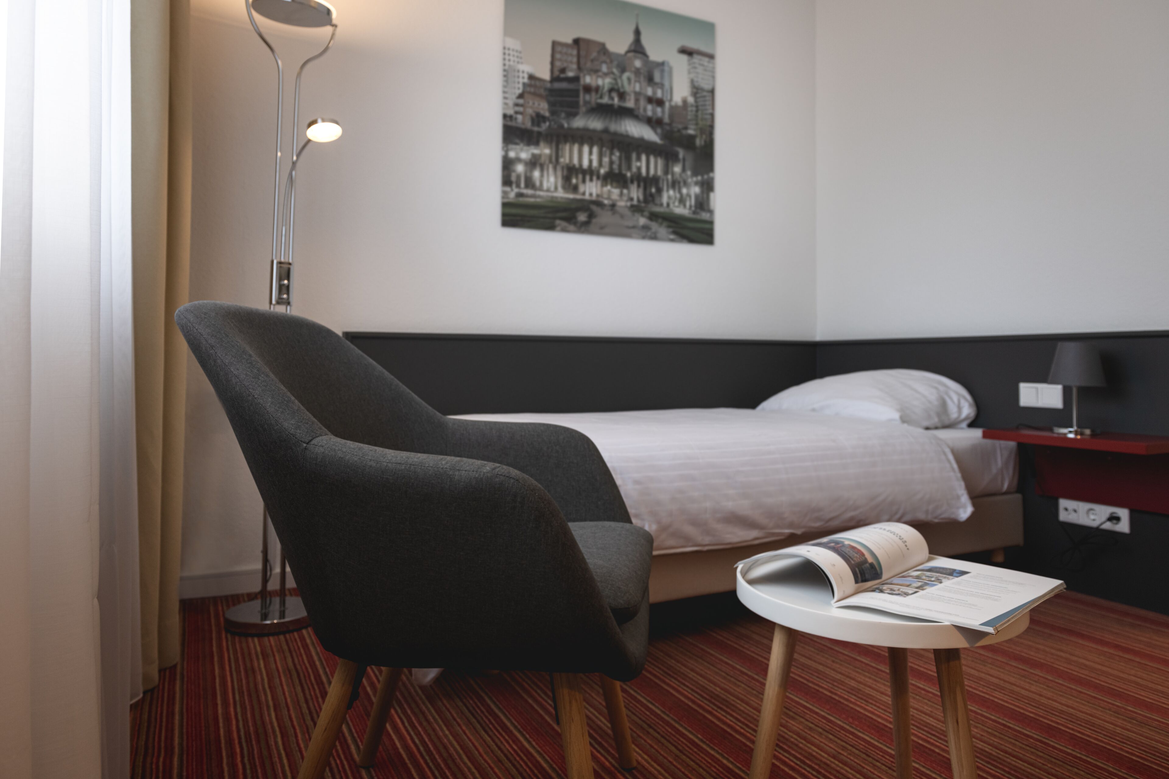 A hotel room with a single bed, a chair, a small table, a lamp, and a framed picture on the wall.