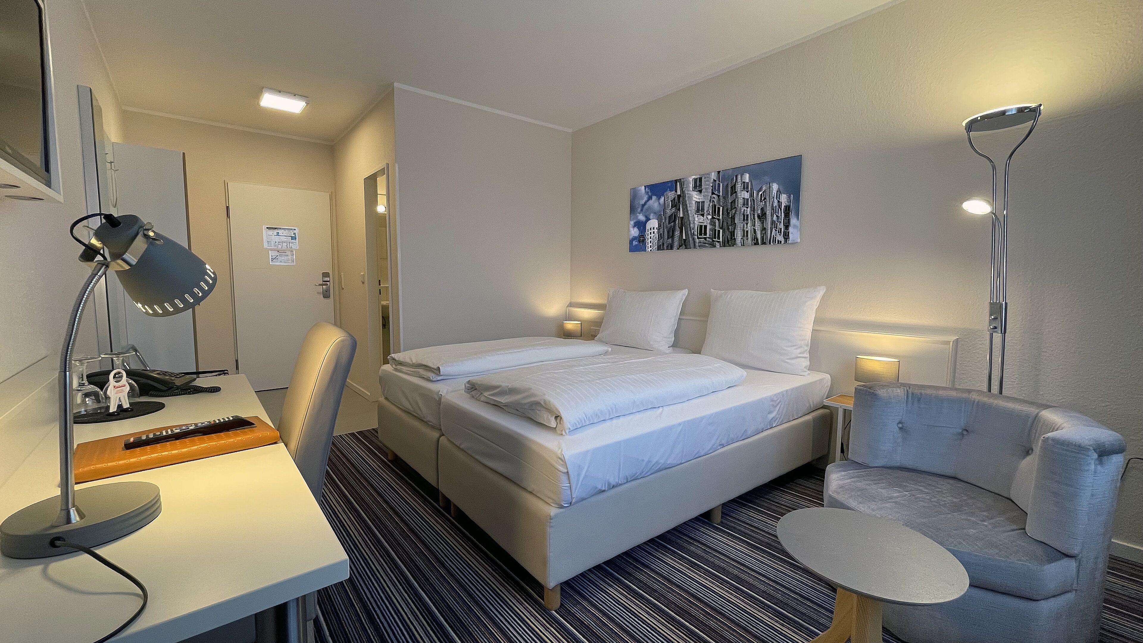 A hotel room with a bed, bedside tables, a desk, a chair, and a floor lamp.
