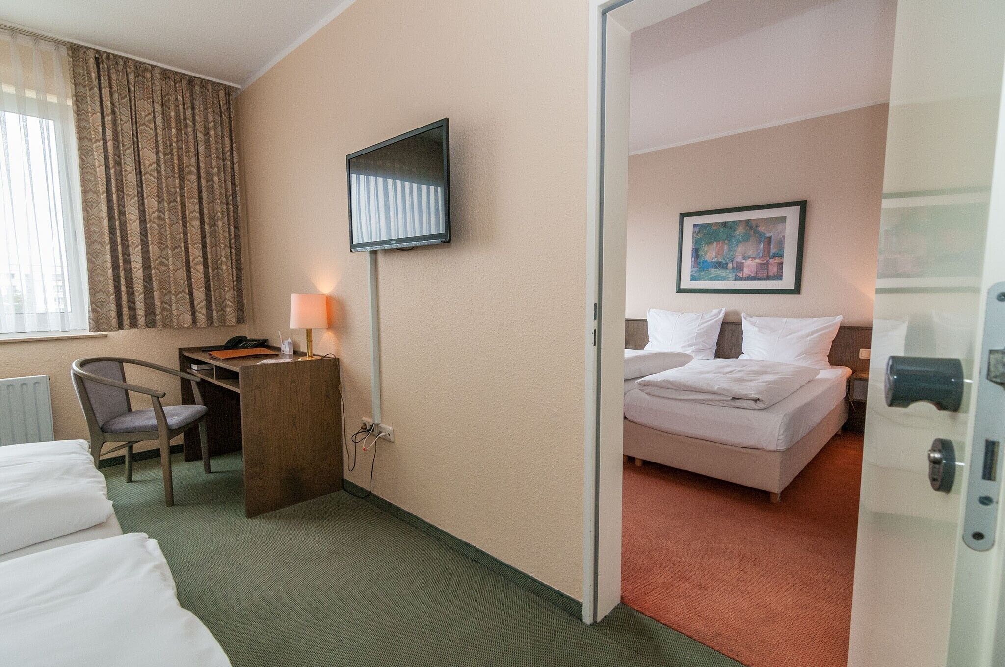 A hotel room with a bed, a desk with a chair, a TV mounted on the wall, and a painting on the wall.