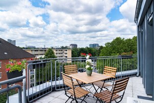 Un balcone con tavolo e sedie, piante in vaso e vista su edifici e alberi.