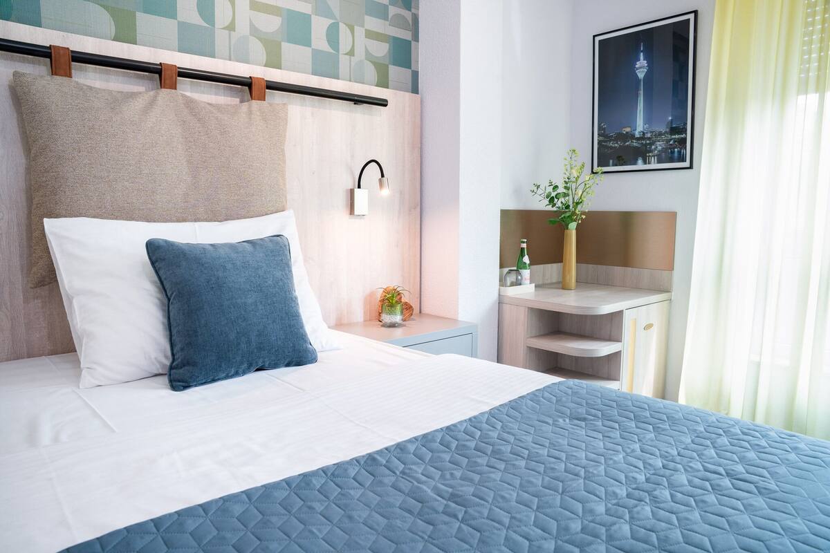 A neatly made bed with a quilted blue cover, a white pillow, and a blue pillow. There is a nightstand with a small plant and a bottle of water. A framed picture of a tower is on the wall, and a window with curtains is visible.