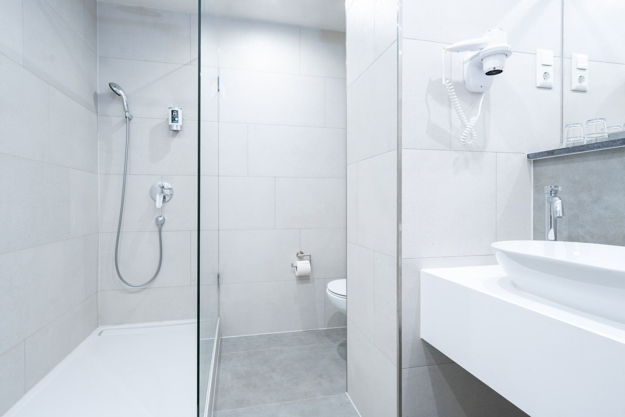 Ein modernes Badezimmer mit einer gläsernen Duschkabine, einem weißen Waschbecken und einem an der Wand befestigten Föhn.