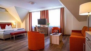 A hotel room with a bed, a desk with a computer, a TV, orange chairs, and a small table with a bottle and glasses. - Hotel Am Stadtpark (Hilden)