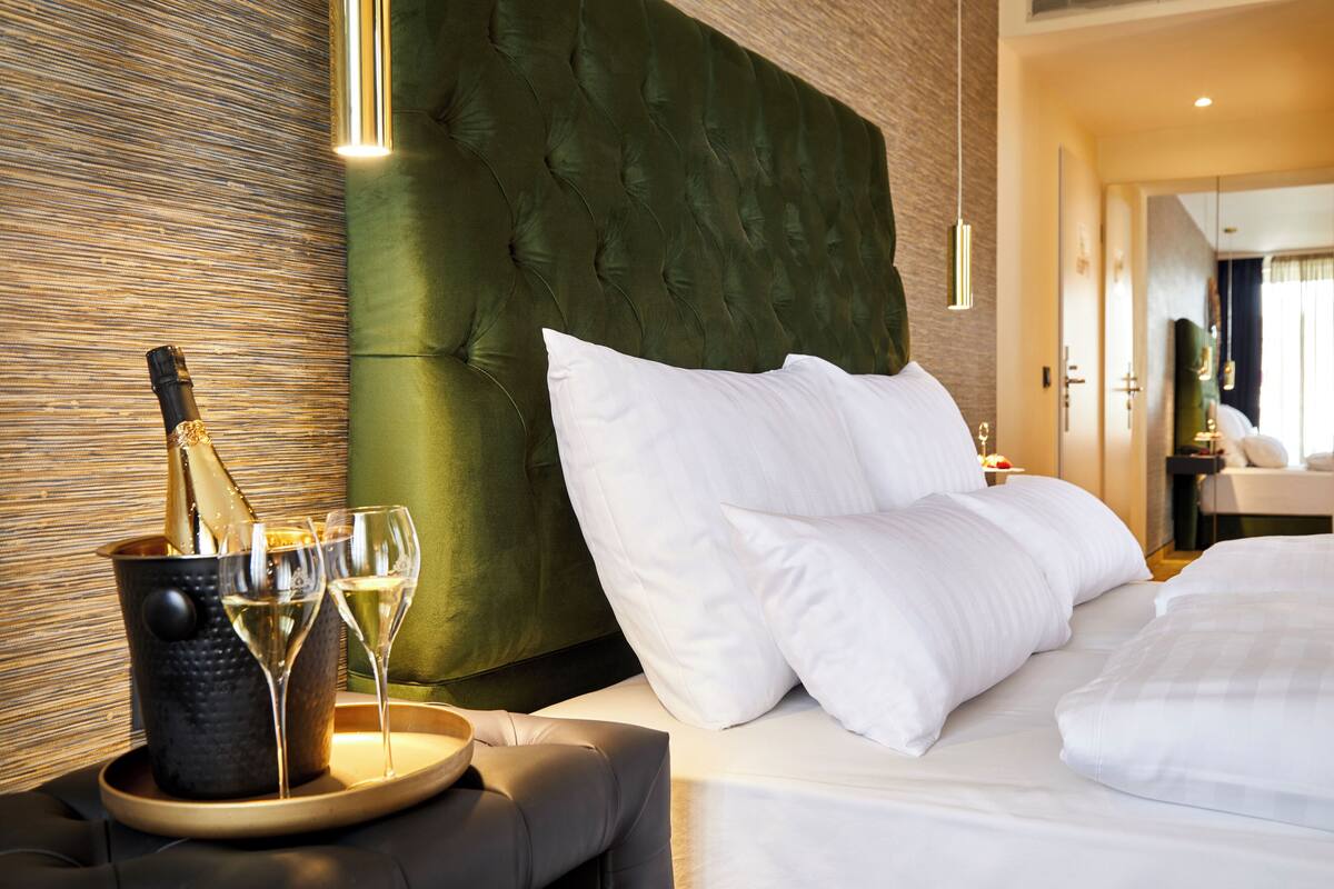 Ein Hotelzimmer mit einer gesteppten Kopfteil, einem Bett mit weißen Leinentüchern, einem Nachttisch mit einem Champagnerkühlbottich und Gläsern sowie einem Holzboden.