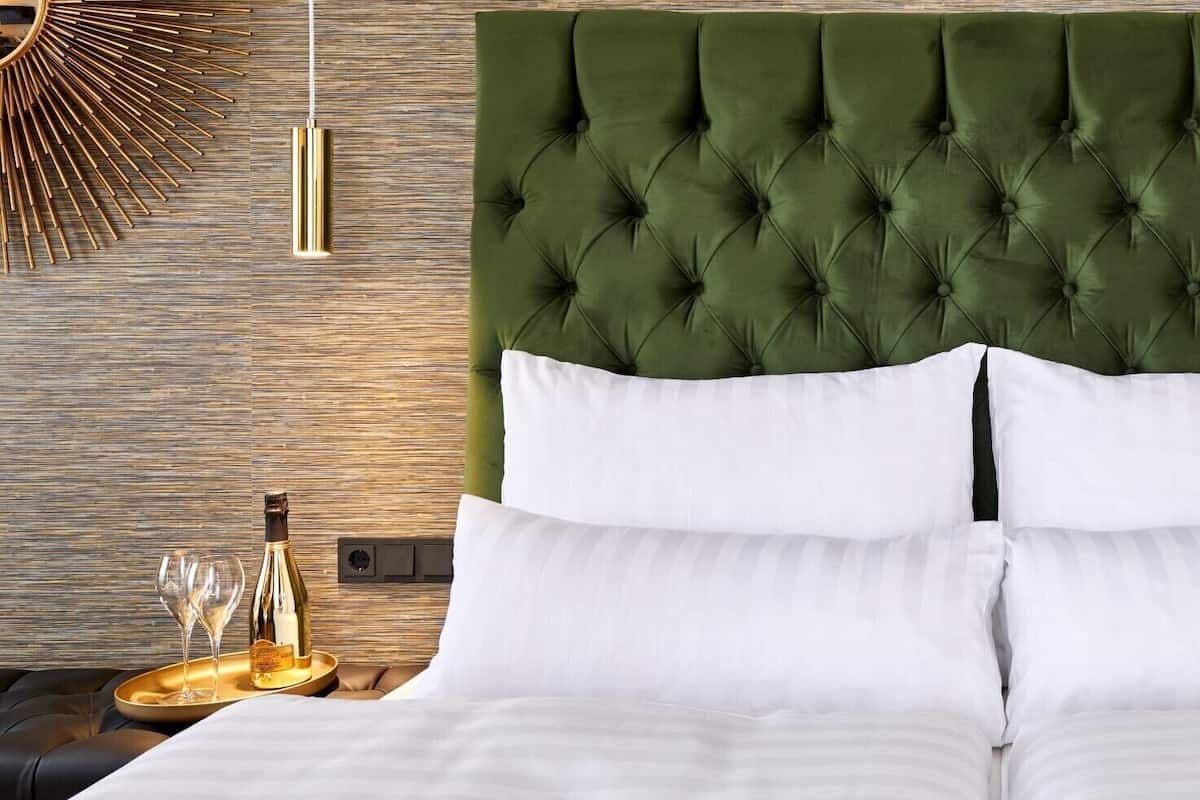 Ein Bett mit einem gesteppten, grünen Kopfteil, weißen Kissen, einem Nachttisch mit Flasche und Gläsern sowie einer kleinen Obstauslage.