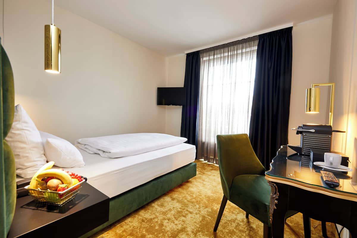 Ein Hotelzimmer mit einem Bett, einem Schreibtisch mit Stuhl, einer Obstschale, einer Fernbedienung und einem an der Wand befestigten Fernseher.