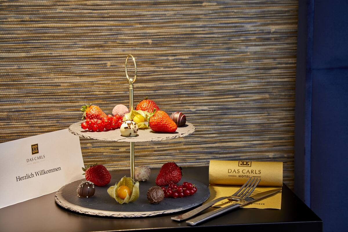 Eine dreistufige Dessertdarbietung mit verschiedenen Früchten und Pralinen, ergänzt durch eine Willkommenskarte und eine Hotelbroschüre.