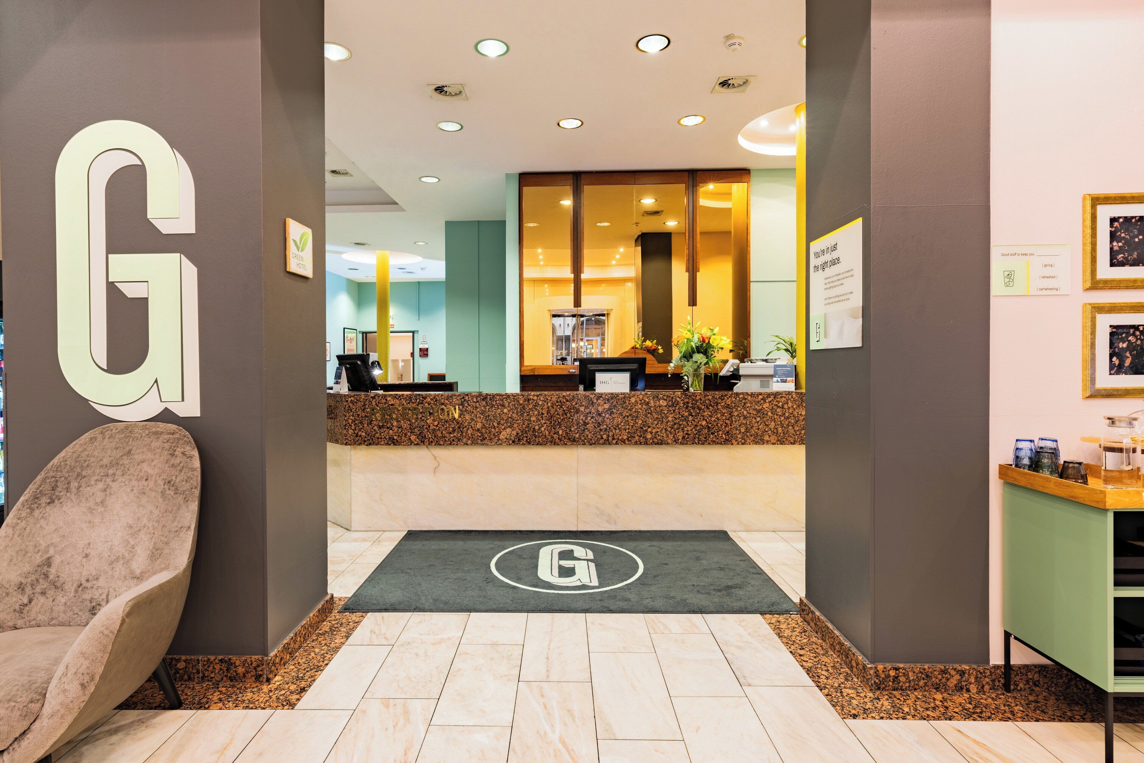 Uma área de recepção com um grande logotipo 'G' na parede, um balcão de recepção, um balcão verde e uma cadeira para o recepcionista.