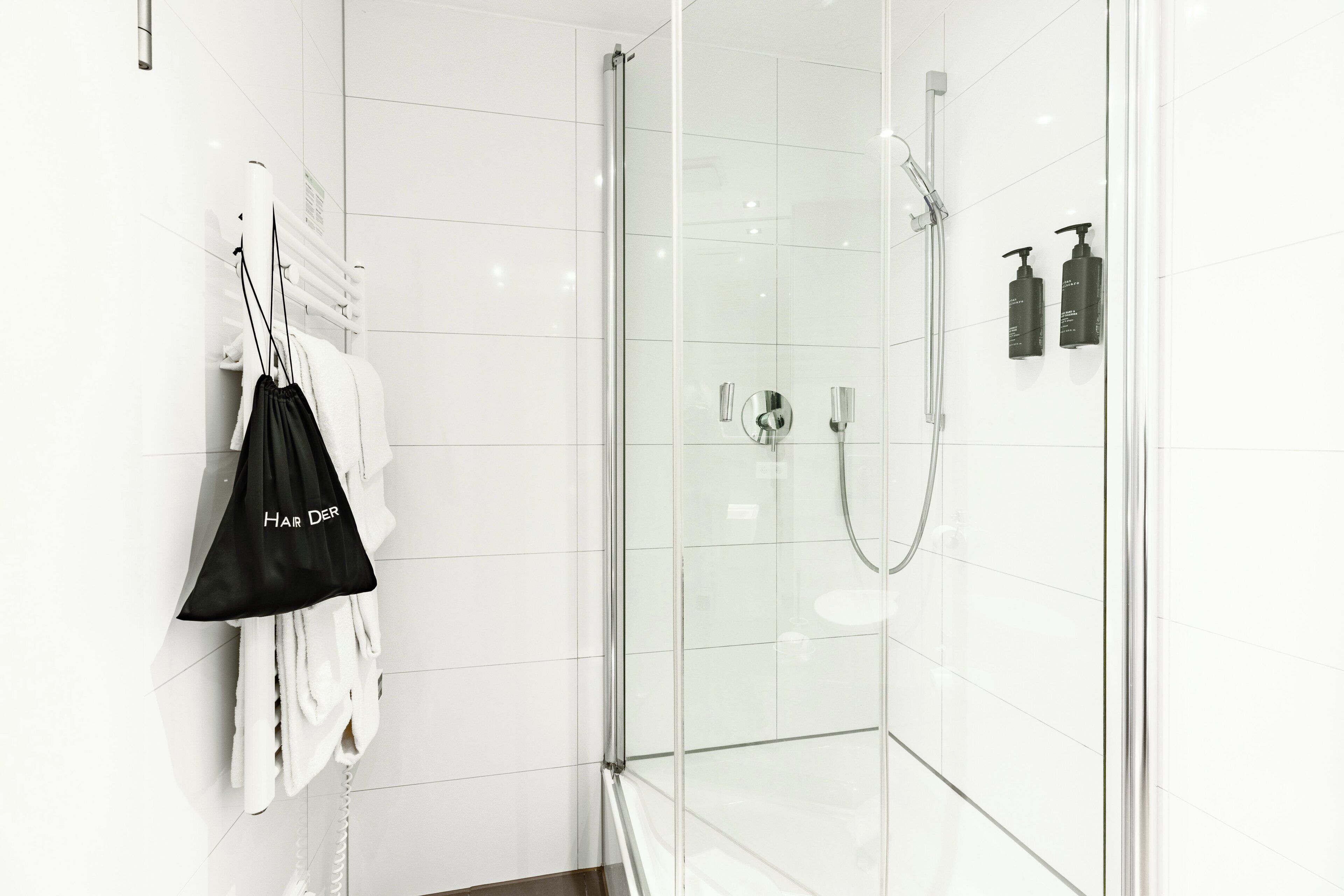 A modern bathroom with a glass-enclosed shower, white tiled walls, and a towel rack with a black bag labeled "Hair Dryer".