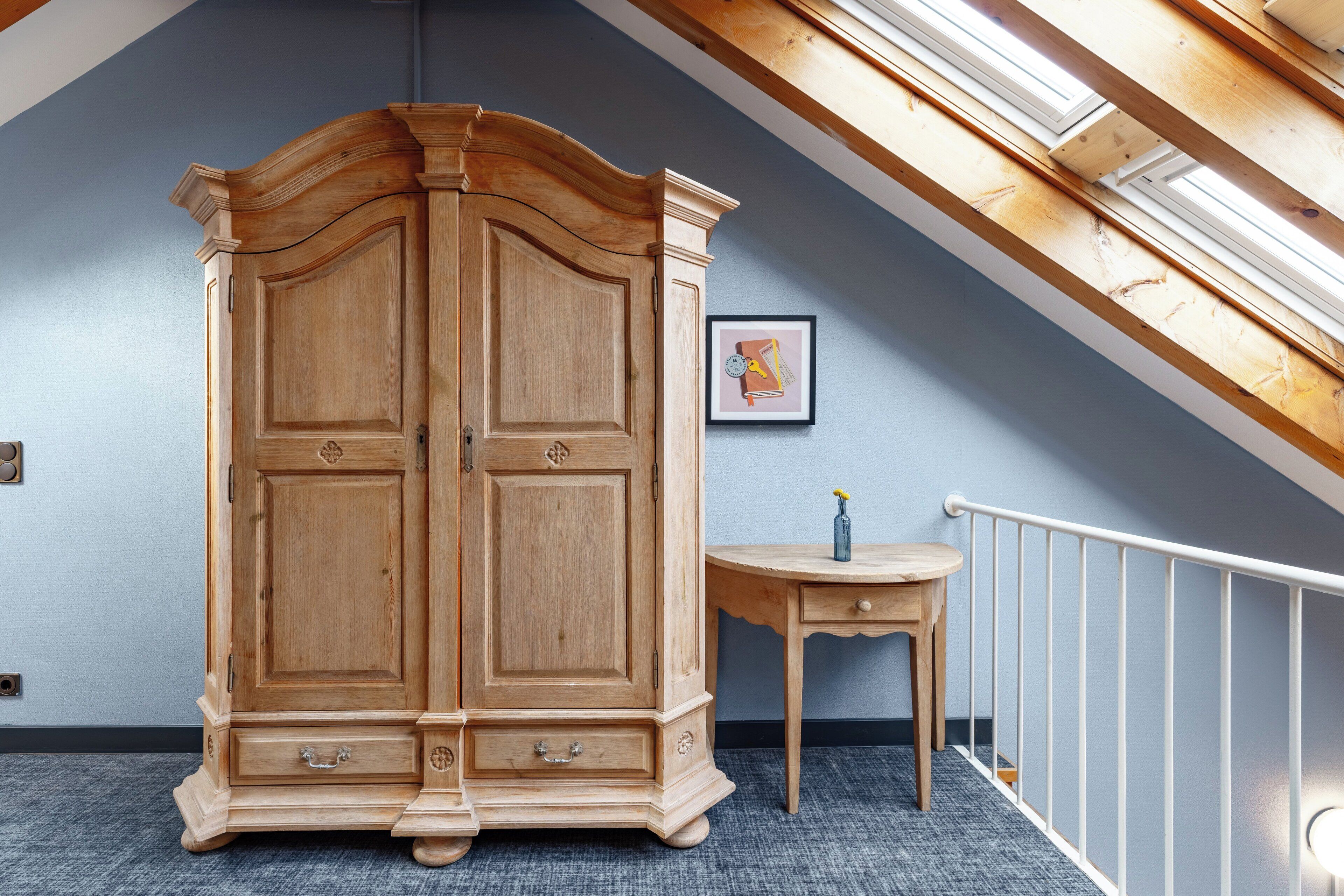 A room with a large wooden wardrobe, a small wooden table, and a framed picture on the wall.