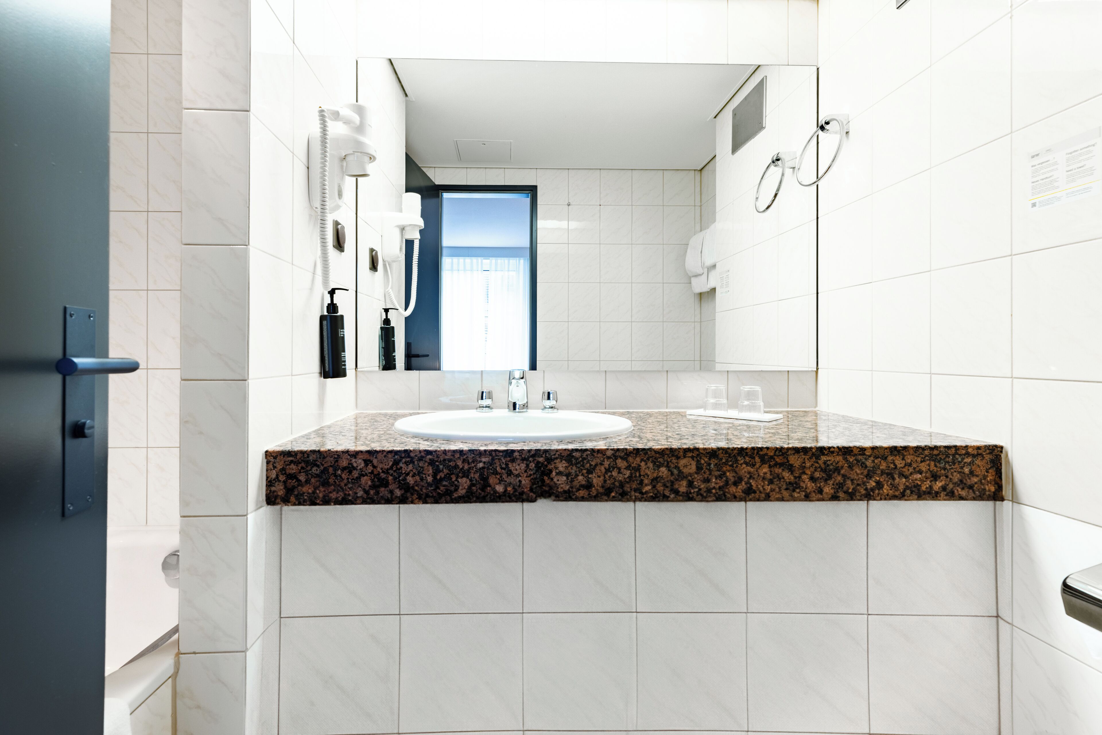 Un baño moderno con lavamanos de granito, azulejos blancos, un espejo y una puerta visible que da a otra habitación.