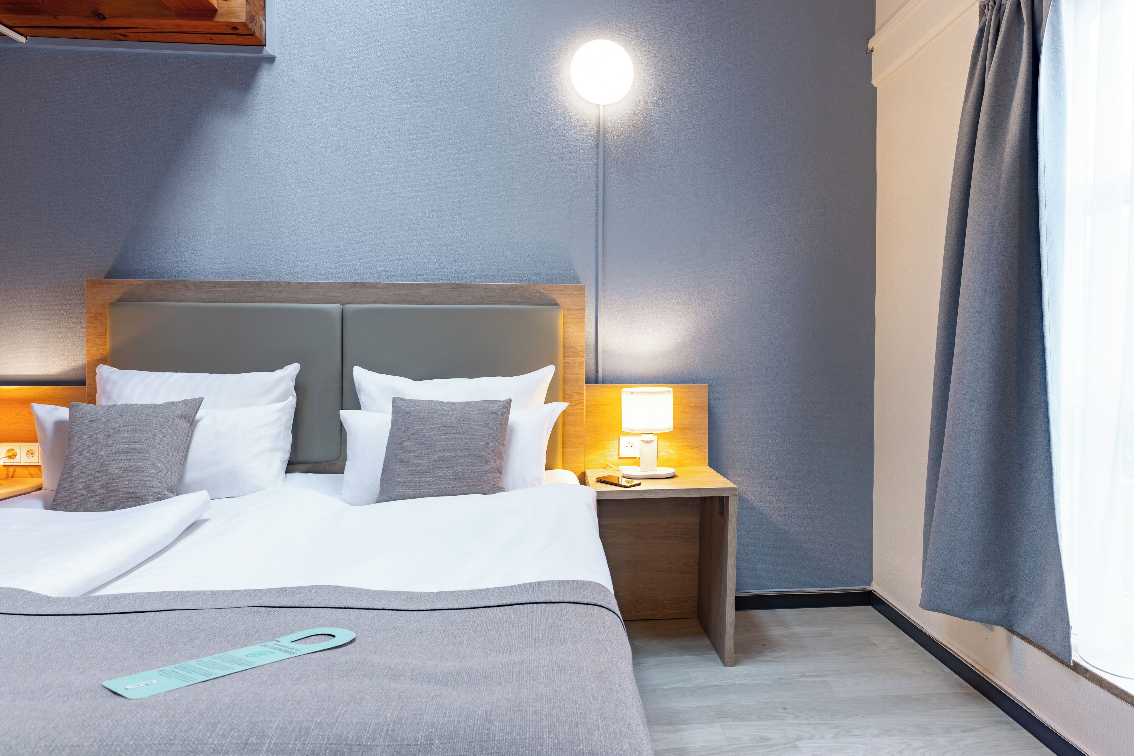 Ein Hotelzimmer mit einem Bett, einem Nachttisch mit Lampe und einem Fenster mit Vorhängen.
