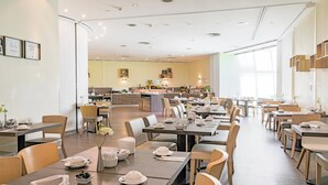 A spacious dining area with multiple tables and chairs, a central kitchen area, and large windows allowing natural light. - Garner Hotel Dusseldorf Main Station by IHG (Düsseldorf)