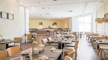 Un'ampia sala da pranzo con diversi tavoli e sedie, una zona cucina centrale e ampie finestre che permettono l'ingresso di luce naturale.