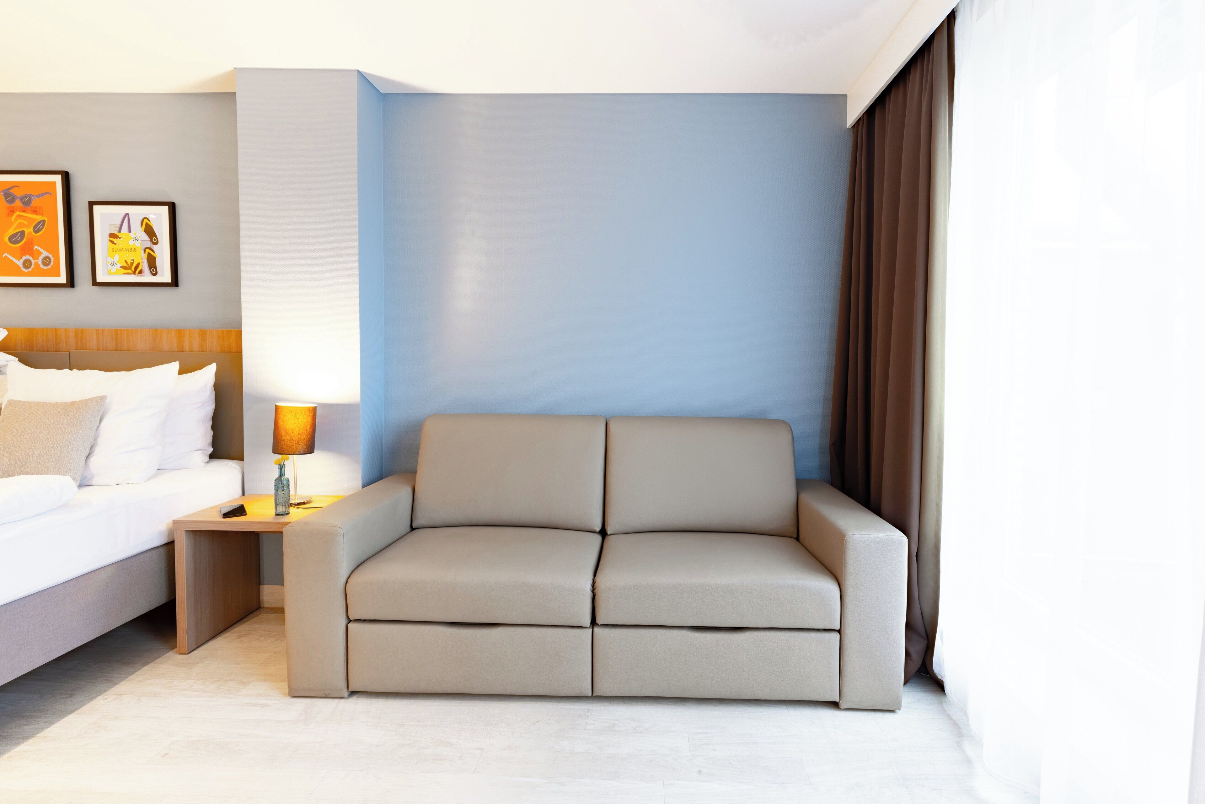 A modern hotel room with a bed, a sofa, a nightstand with a lamp, and framed artwork on the wall.