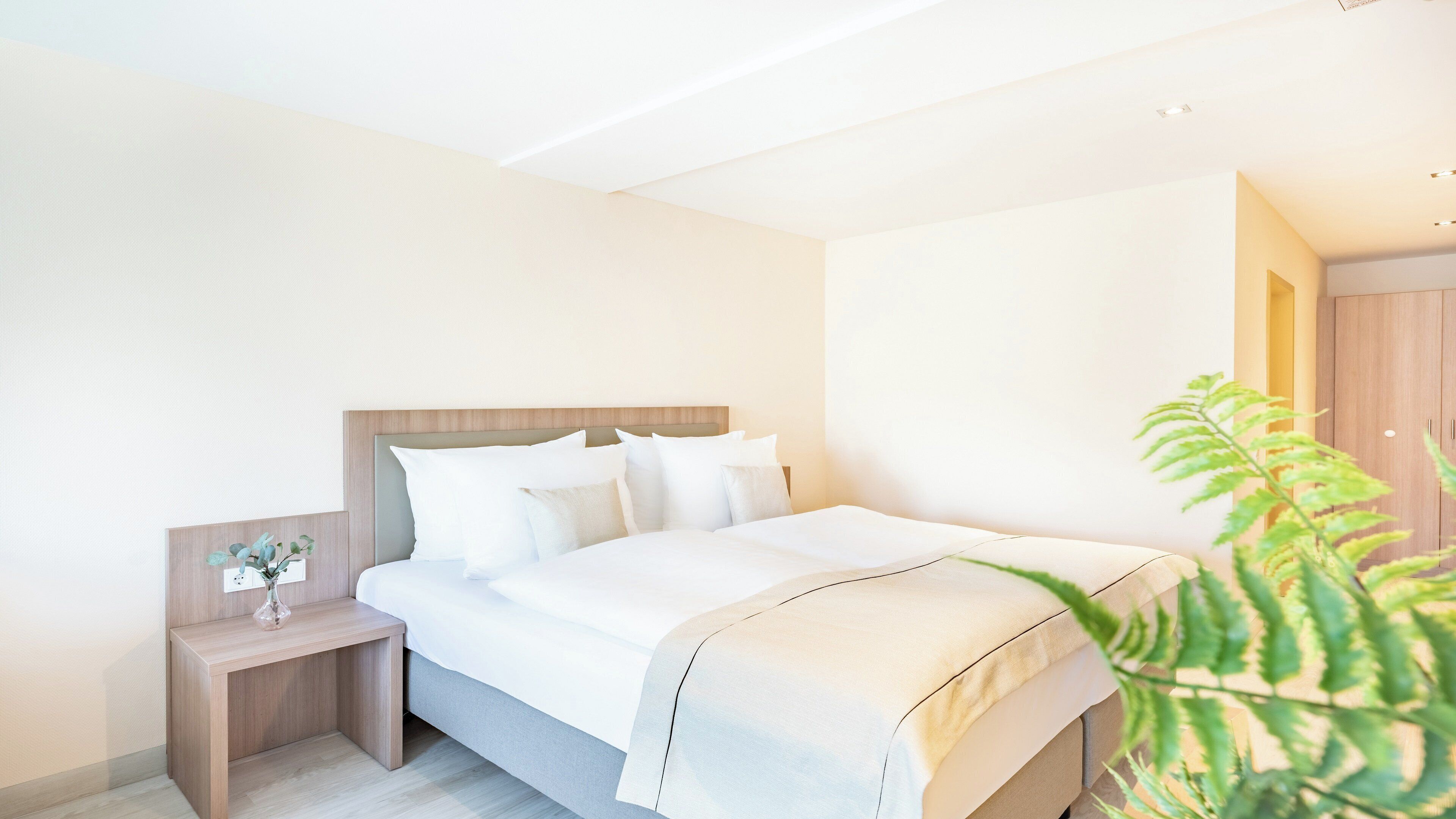 A modern hotel room with a large bed, a bedside table with a vase, and a wooden wardrobe.