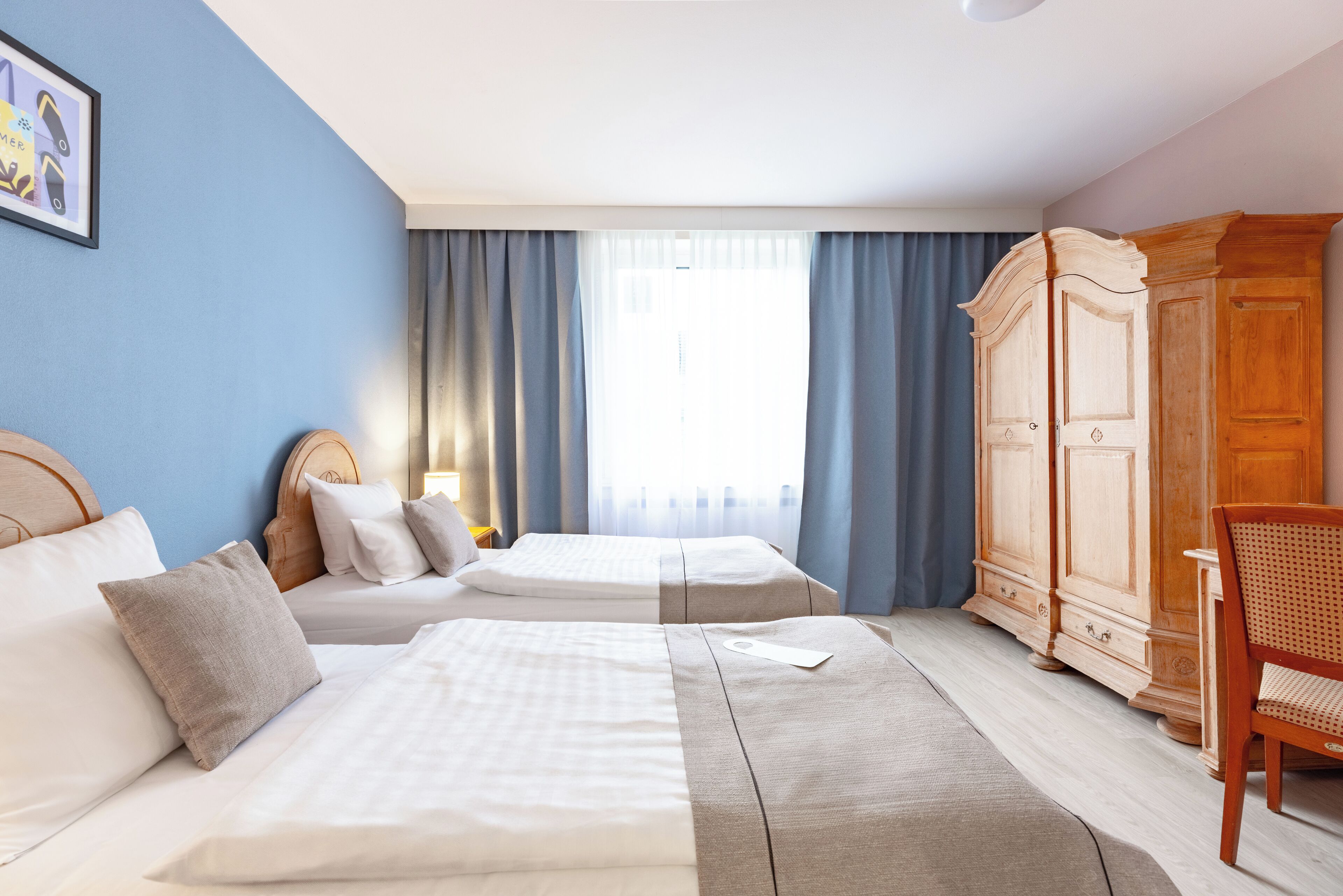 Habitació d'hotel amb dos llits, un armari de fusta, una cadira i una finestra gran amb cortines.