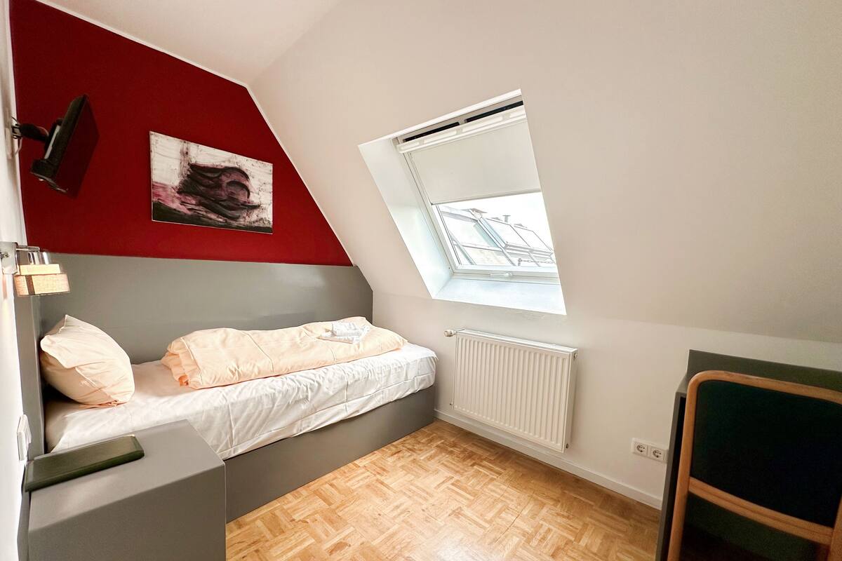 Una habitación pequeña con techo inclinado, una cama, un radiador, una ventana y un cuadro en la pared.