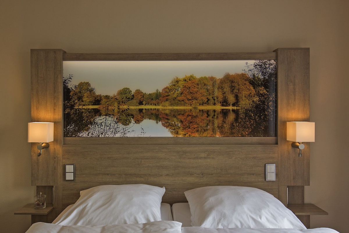 Ein Hotelzimmer mit einem großen Bett, zwei Nachttischlampen und einem Wandbild, das einen von Bäumen umgebenen See zeigt.