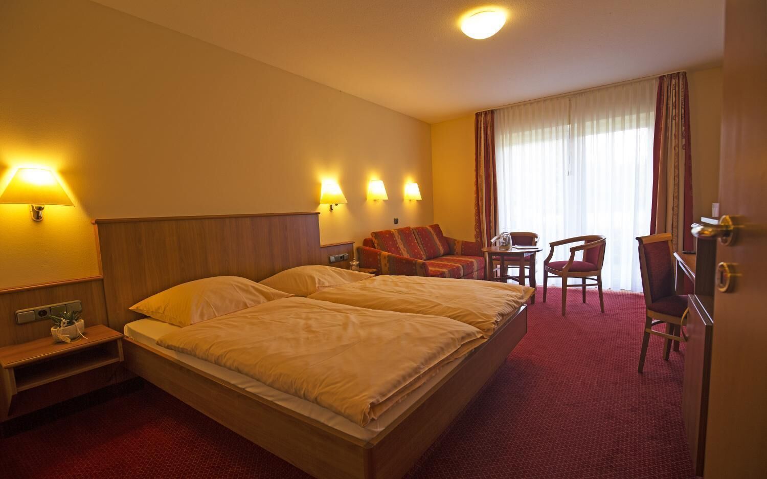 A hotel room with a large bed, two bedside tables, a desk with a chair, a sofa, and a large window with curtains.