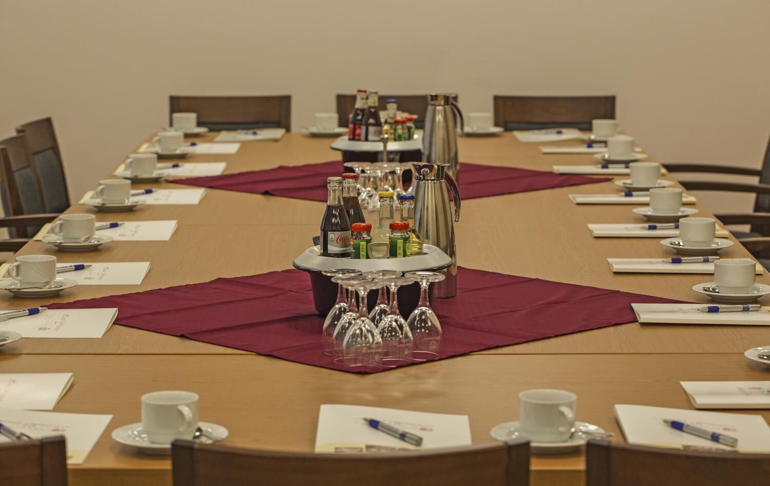 A conference room with a long table set for a meeting, equipped with coffee cups, saucers, and a beverage station with bottles and glasses.