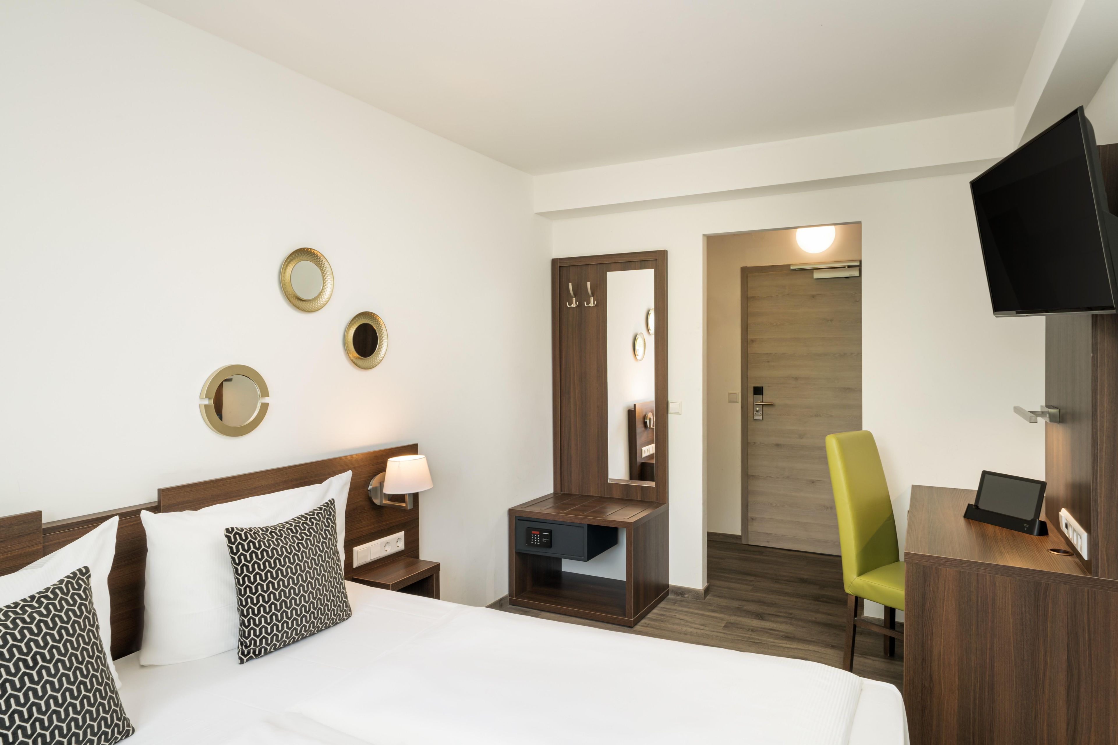 A hotel room with a bed, bedside table, lamp, mirror, and a desk with a chair.
