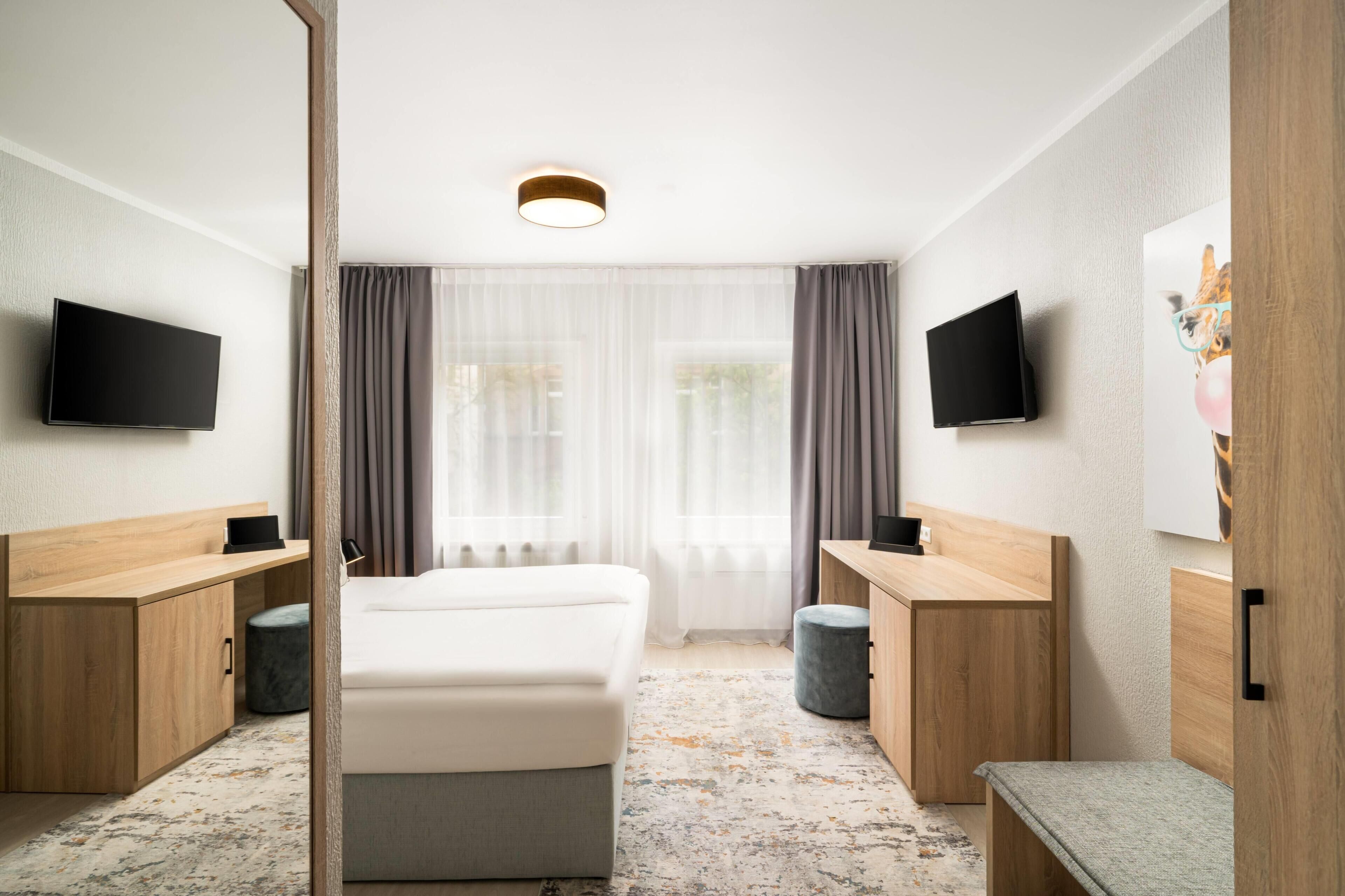 A hotel room with a bed, two bedside tables, a desk, a chair, a flat-screen TV on each wall, and a large window with curtains.